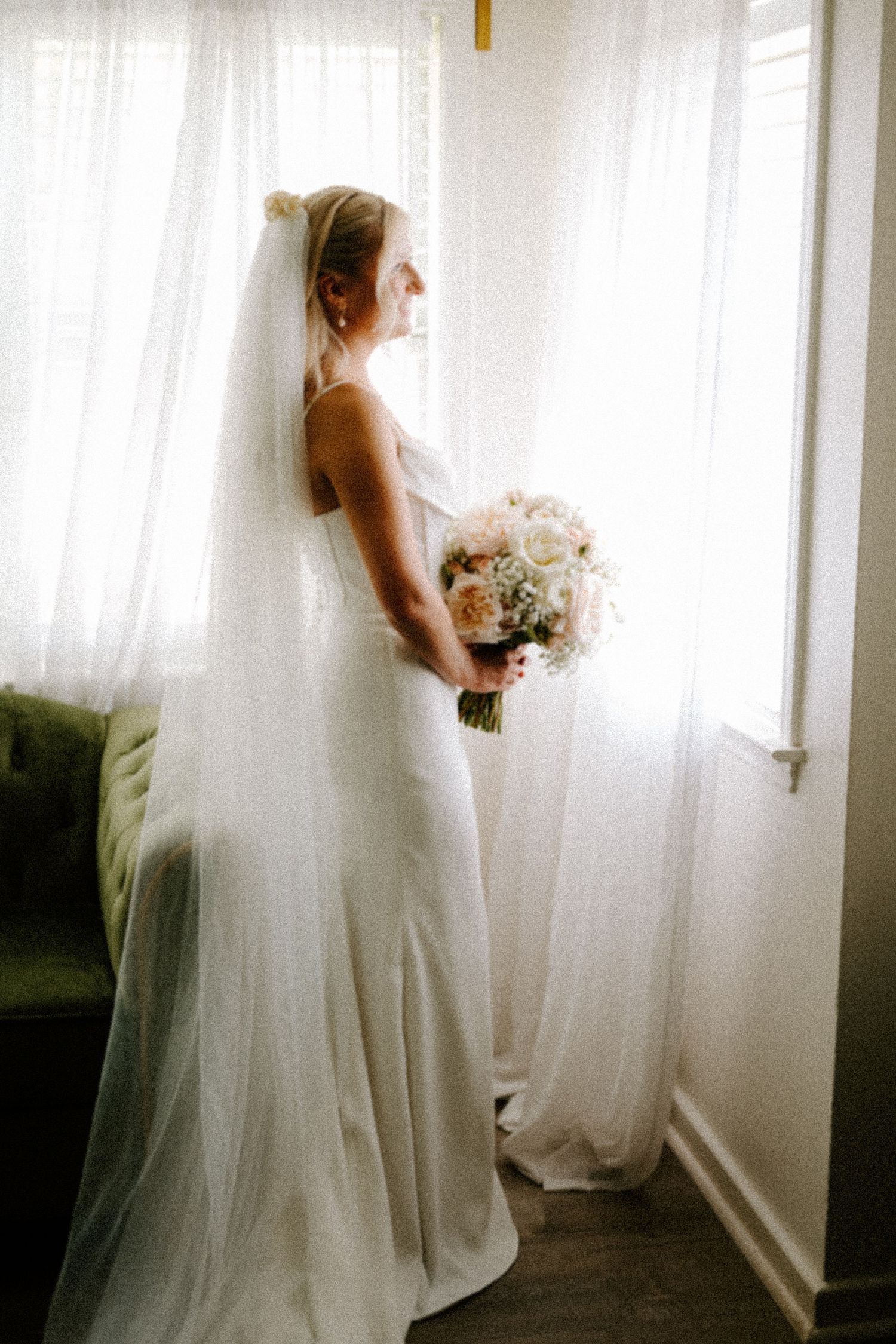 Ethereal bridal silhouettes by window in white room with flowing veil and bouquet.