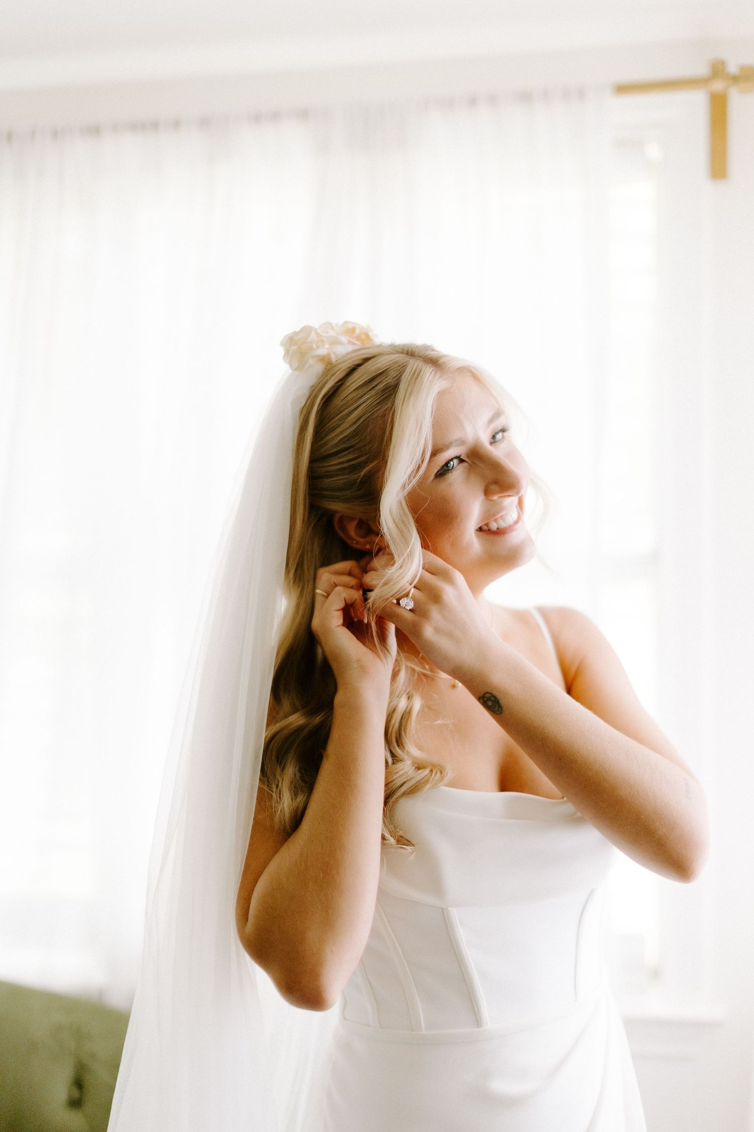 A bride in a white gown puts on earrings while standing by sheer curtains.