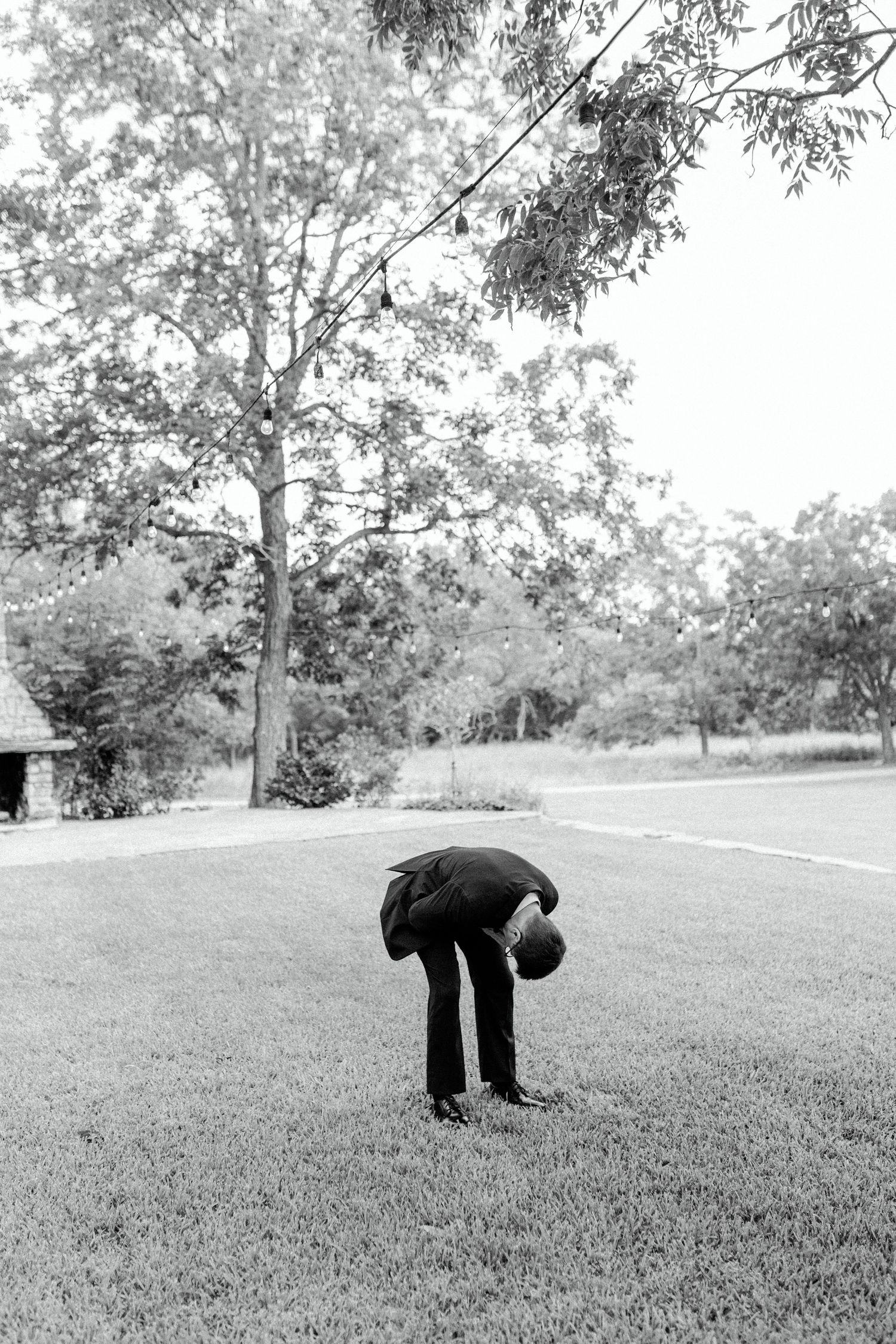 A person in dark clothing bends down to examine something on a grassy lawn with trees in the background.