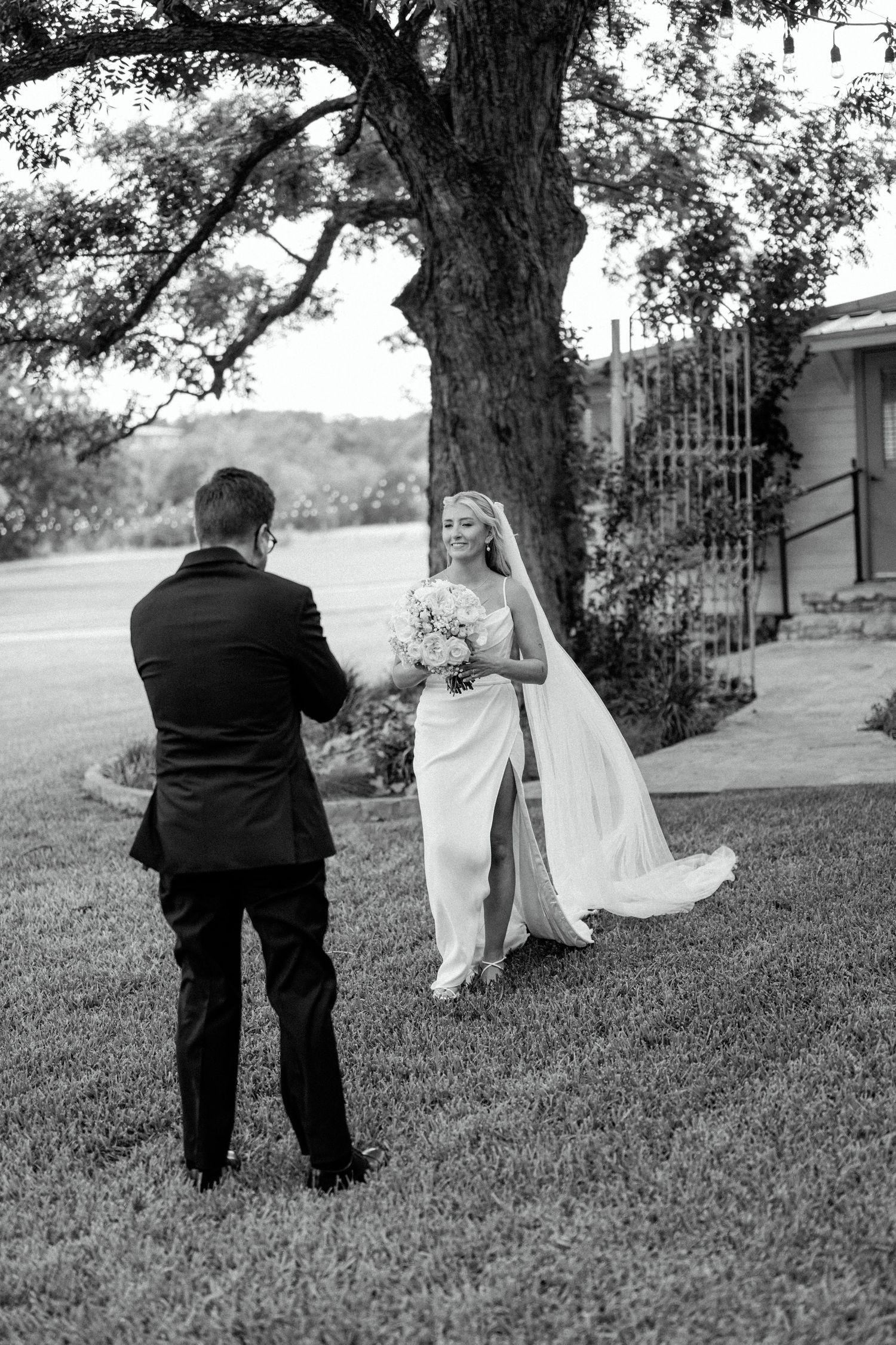 A black and white wedding photo captures a couple during their first look moment under a large tree.