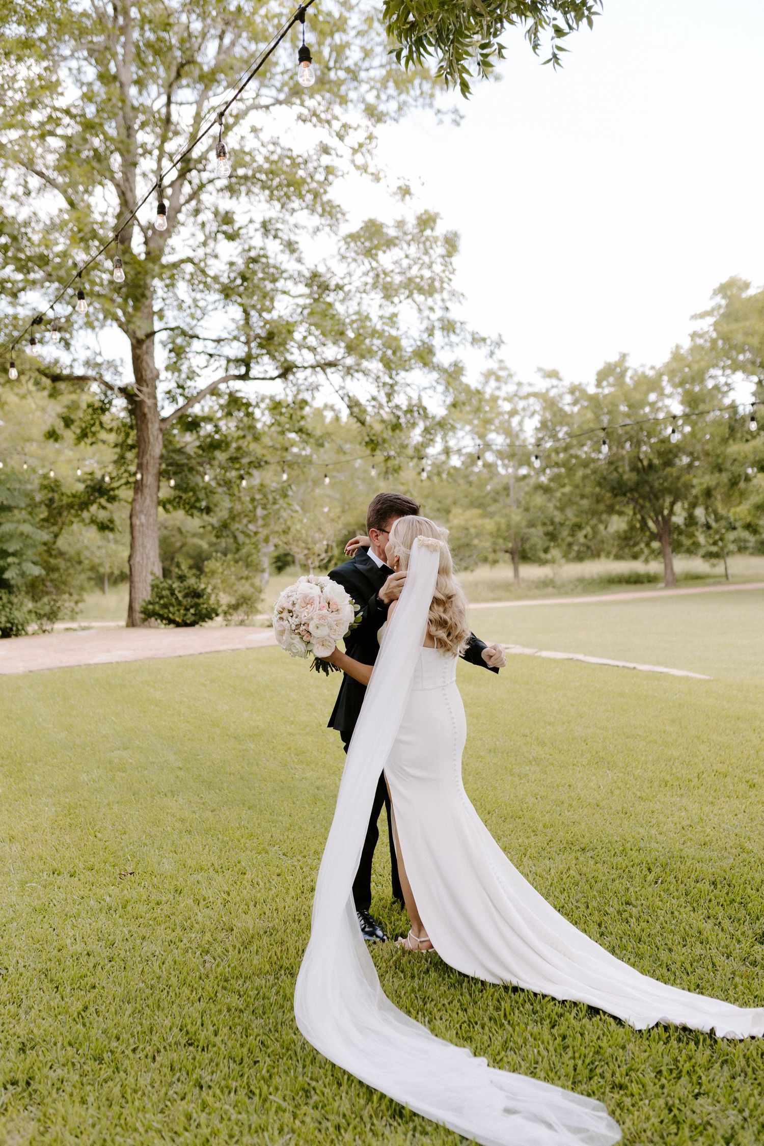 A couple embraces in a romantic moment on a grassy lawn with trees, the bride's long white train flowing behind her.