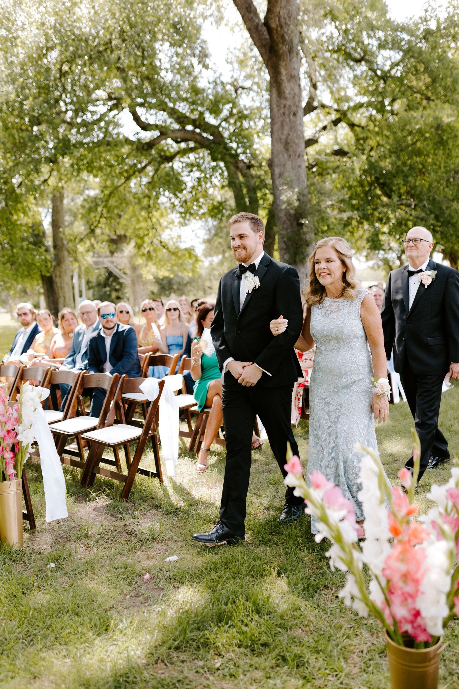 Outdoor wedding ceremony setup with wooden chairs and floral decorations under oak trees.