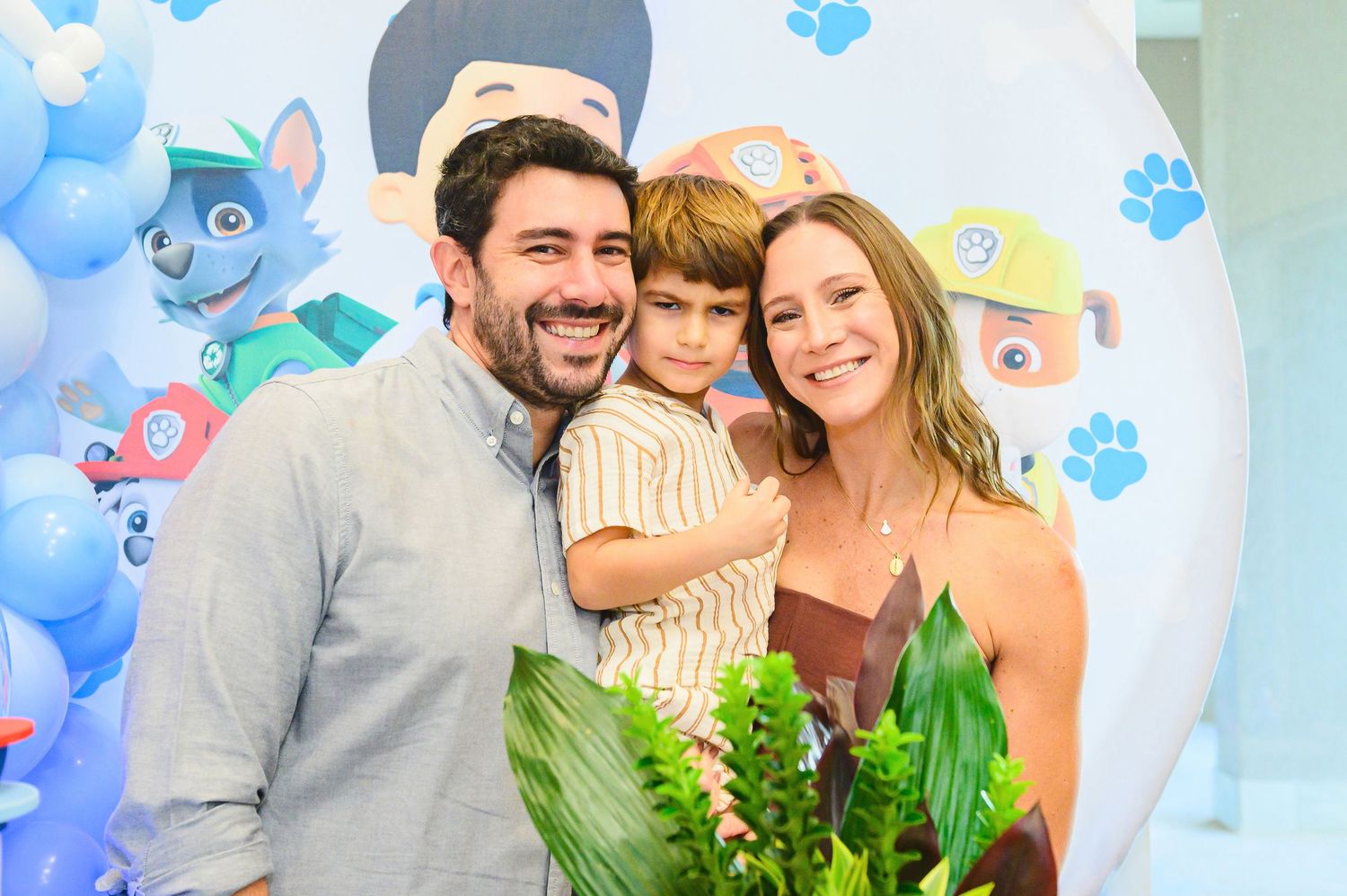 A happy couple stands with their toddler at a party with blue balloon decorations and tropical leaf decorations.