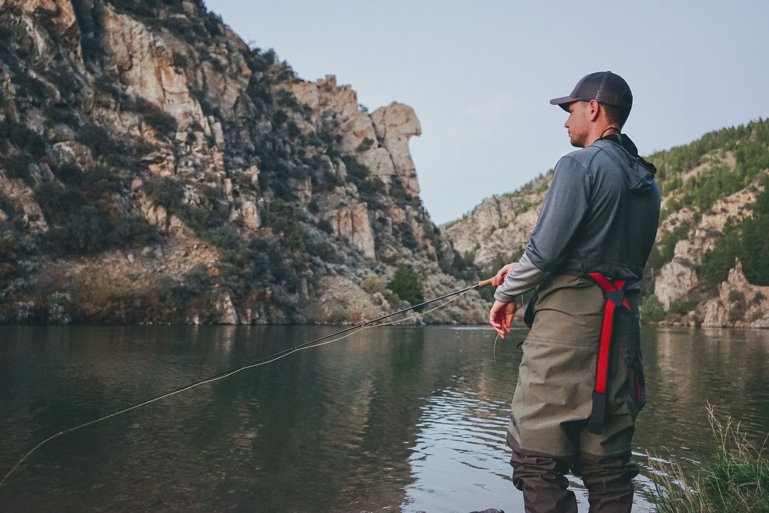 Fly fisherman casting in mountain river while wearing waders and gray jacket.