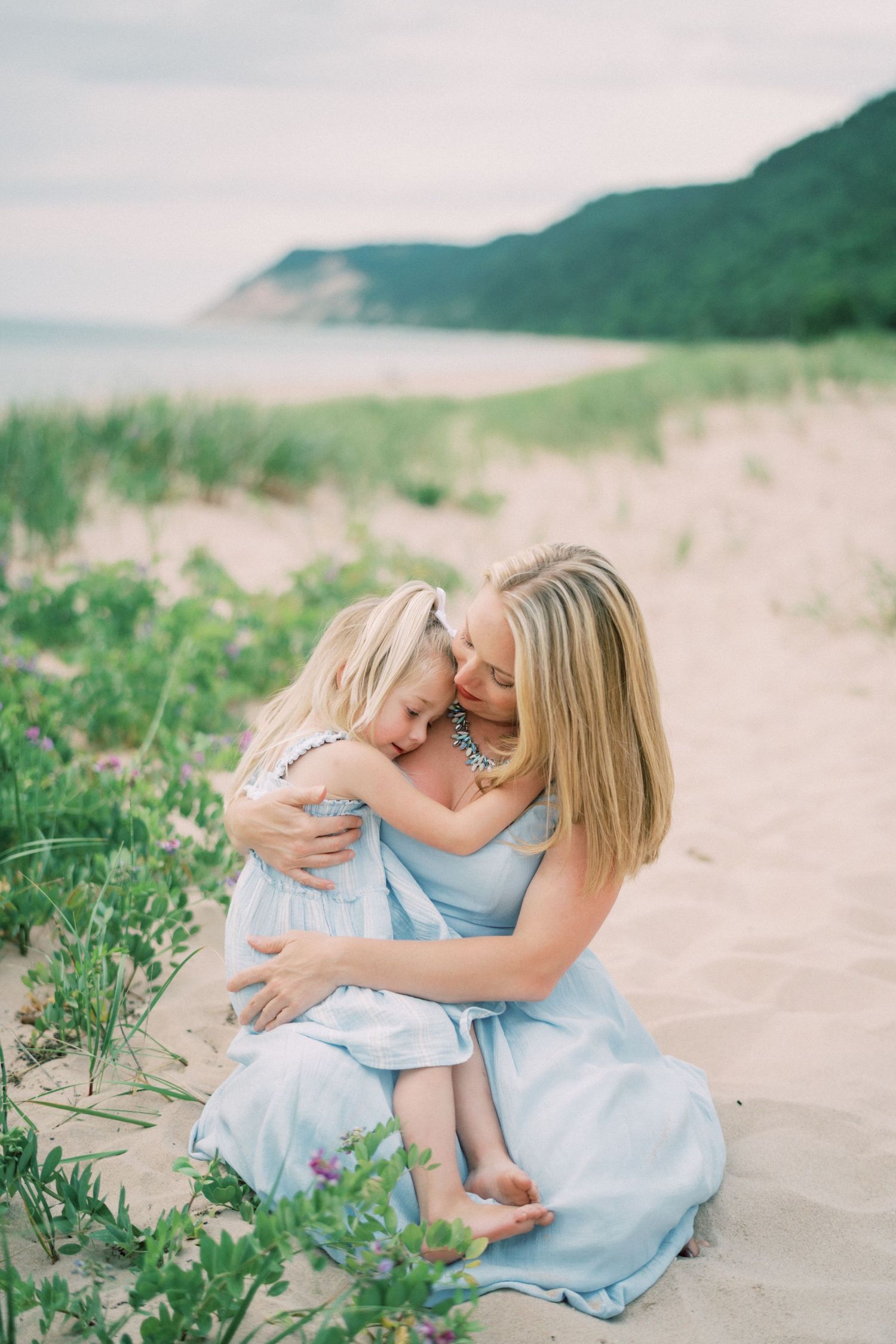 A loving embrace shared on a sandy beach path with green coastal hills and vegetation in the background.