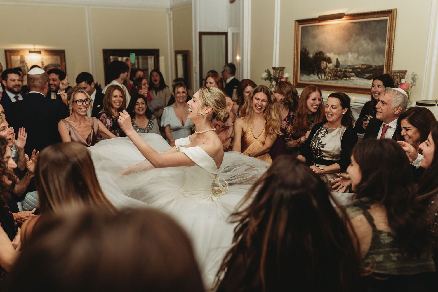 Dance floor filled with wedding guests celebrating at an elegant reception venue with classic chandeliers glowing above.
