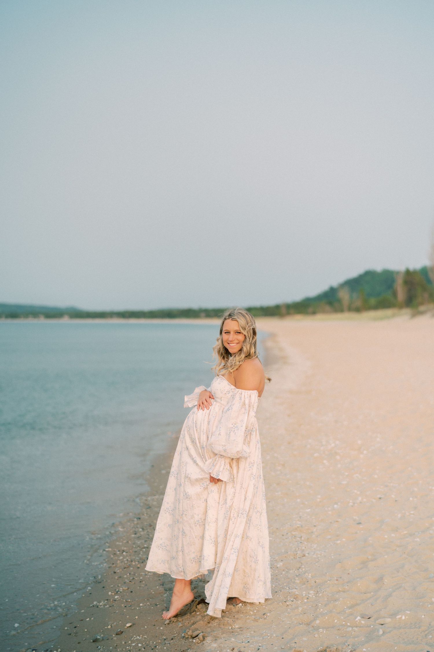 A pregnant woman in a flowing white dress walks barefoot along a peaceful beach shoreline at sunset.