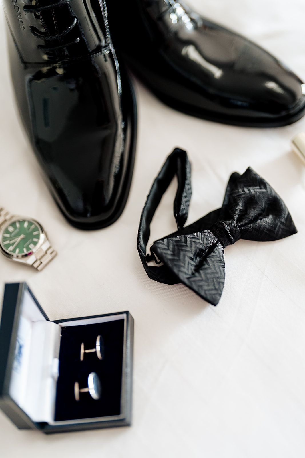 A groom's wedding accessories including shiny black dress shoes, a patterned bow tie, cufflinks in a box, and a wristwatch.