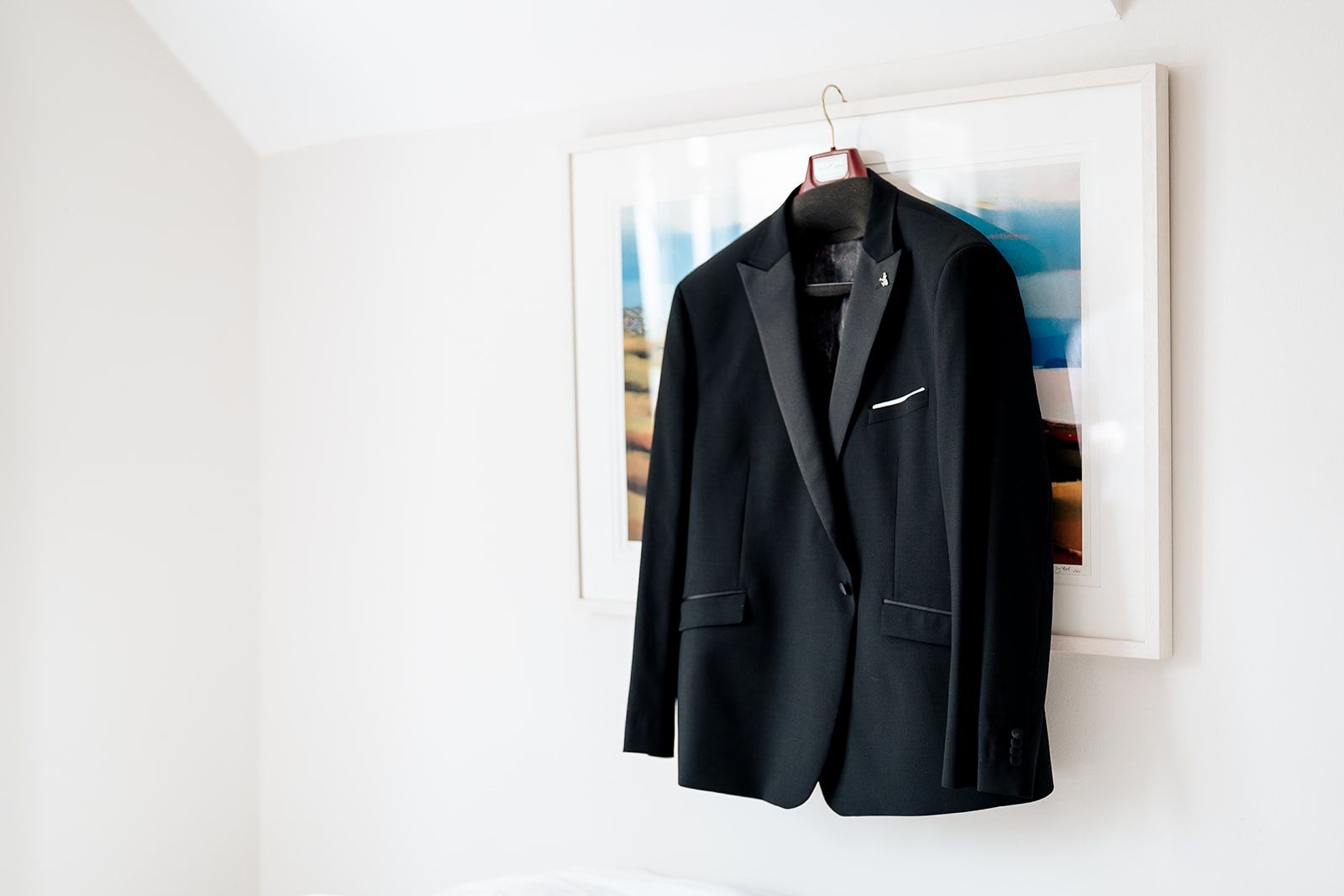 A black formal suit jacket hangs on a hanger against a white wall with modern artwork in the background.