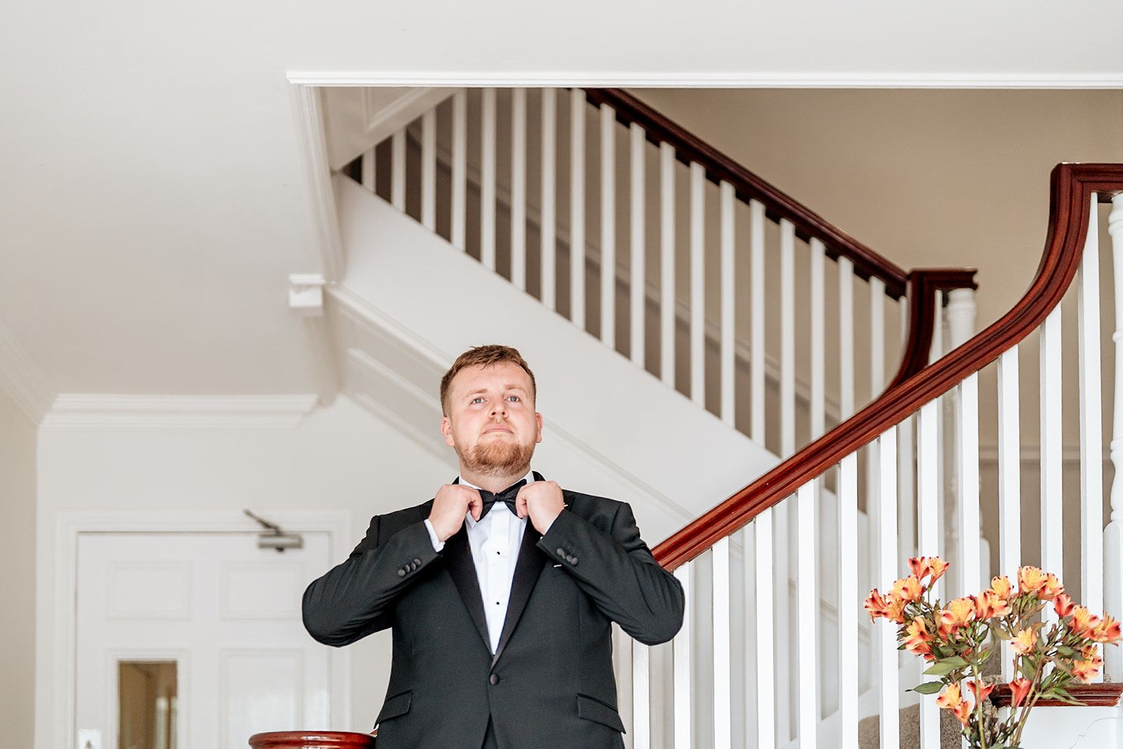 A person in a black tuxedo adjusts their pink bow tie while standing at the bottom of an elegant white staircase.