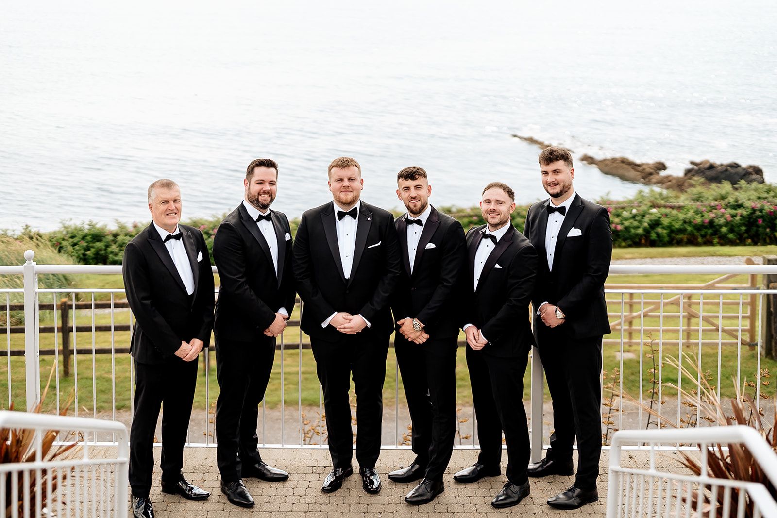 Groom and groomsmen in black tuxedos stand together on a scenic beachfront deck with coastal views in background.