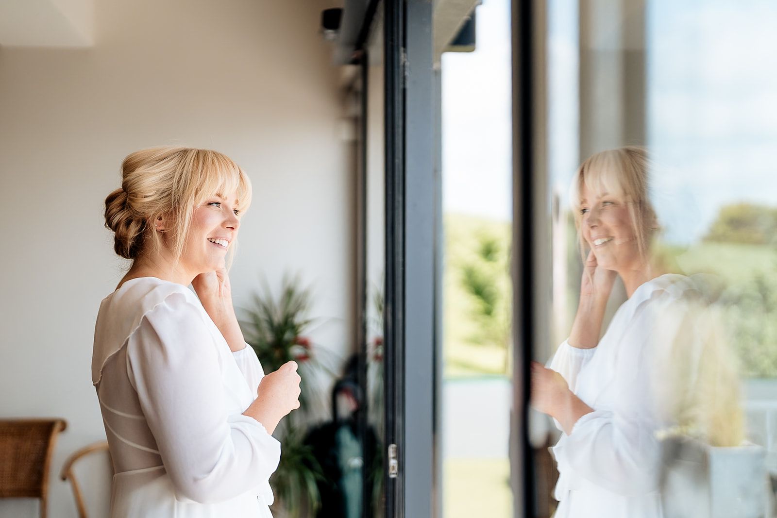 A bride in a classic white wedding gown looks thoughtfully out a window in an elegant indoor setting.
