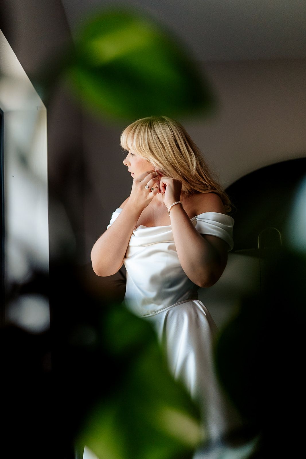 Person in a white dress standing with hands covering their face in dim lighting with green foliage visible in background.