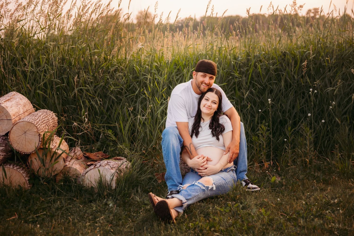 Couple sitting together on grass near stacked logs during golden hour sunset with tall grass in background.