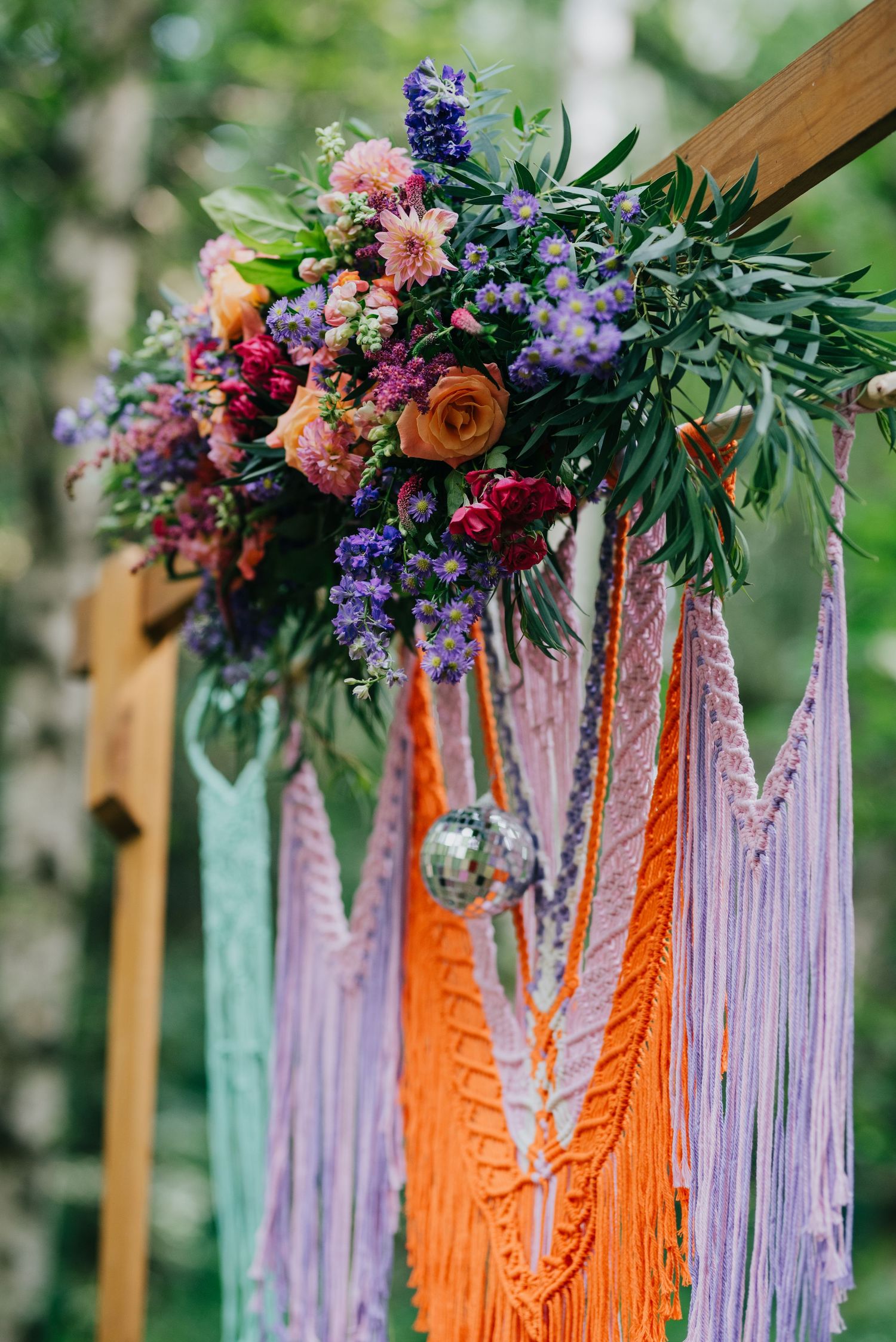 Colorful floral arrangement with purple and pink flowers adorned with hanging macrame in orange and lavender tones.