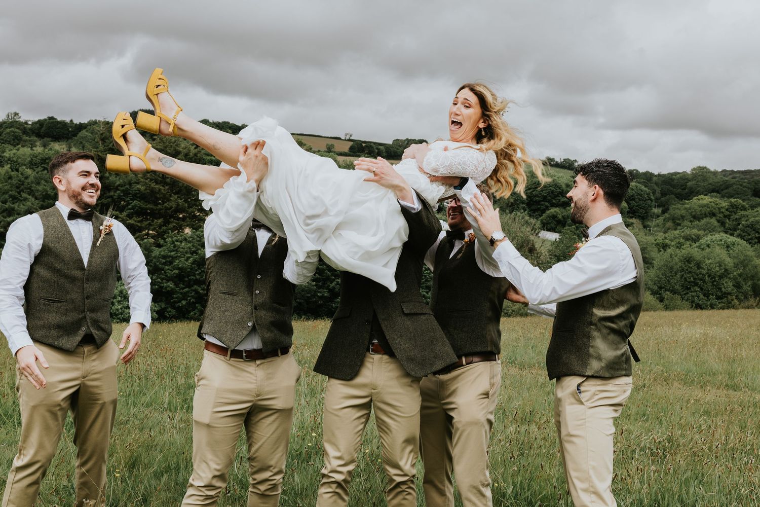 Fun wedding party in matching tan pants and green vests playfully lift someone wearing a white dress in a grassy field.