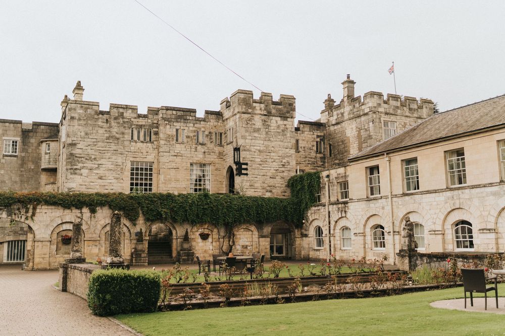 Historic stone castle courtyard with ivy-covered walls, manicured hedges, and elegant architectural features in muted tones.