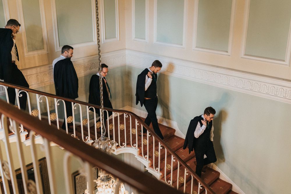 Groomsmen in black tuxedos walking down an elegant curved white staircase with wooden bannister.