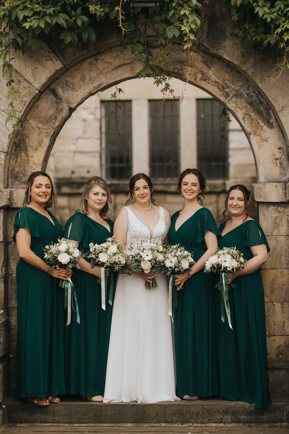 Wedding party in emerald green dresses and white bouquets poses under stone archway during celebration.