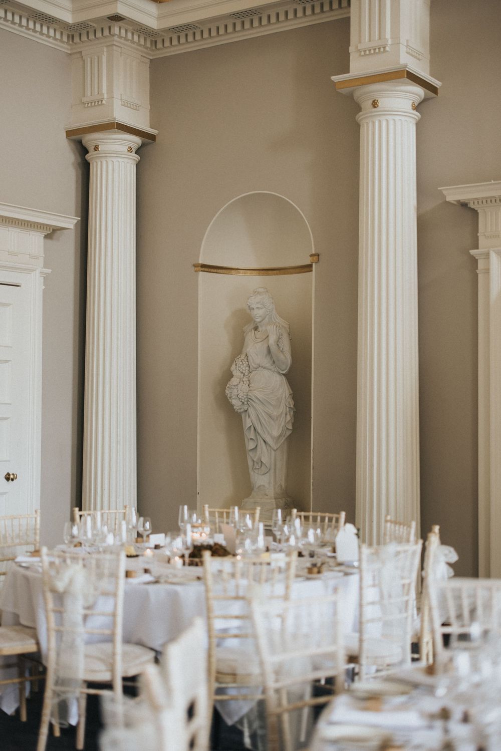 Elegant banquet hall set with white tables and chairs featuring classical architectural columns and statues.