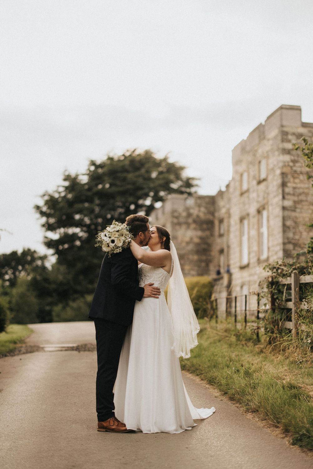A couple in wedding attire embraces on a country path near stone buildings at their rustic outdoor wedding ceremony.