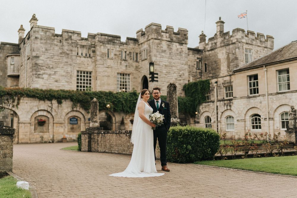 A wedding couple stands in front of a historic stone castle with ivy-covered walls and manicured grounds.