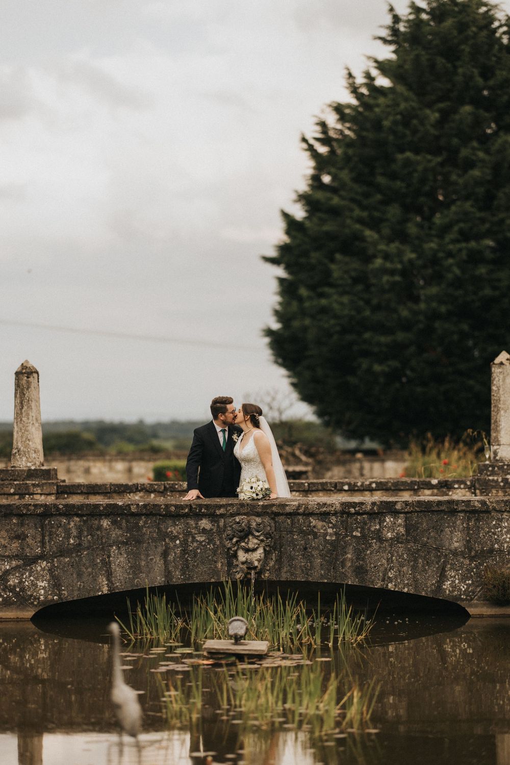 A romantic wedding couple stands on an old stone bridge over calm water with a large evergreen tree in the background.