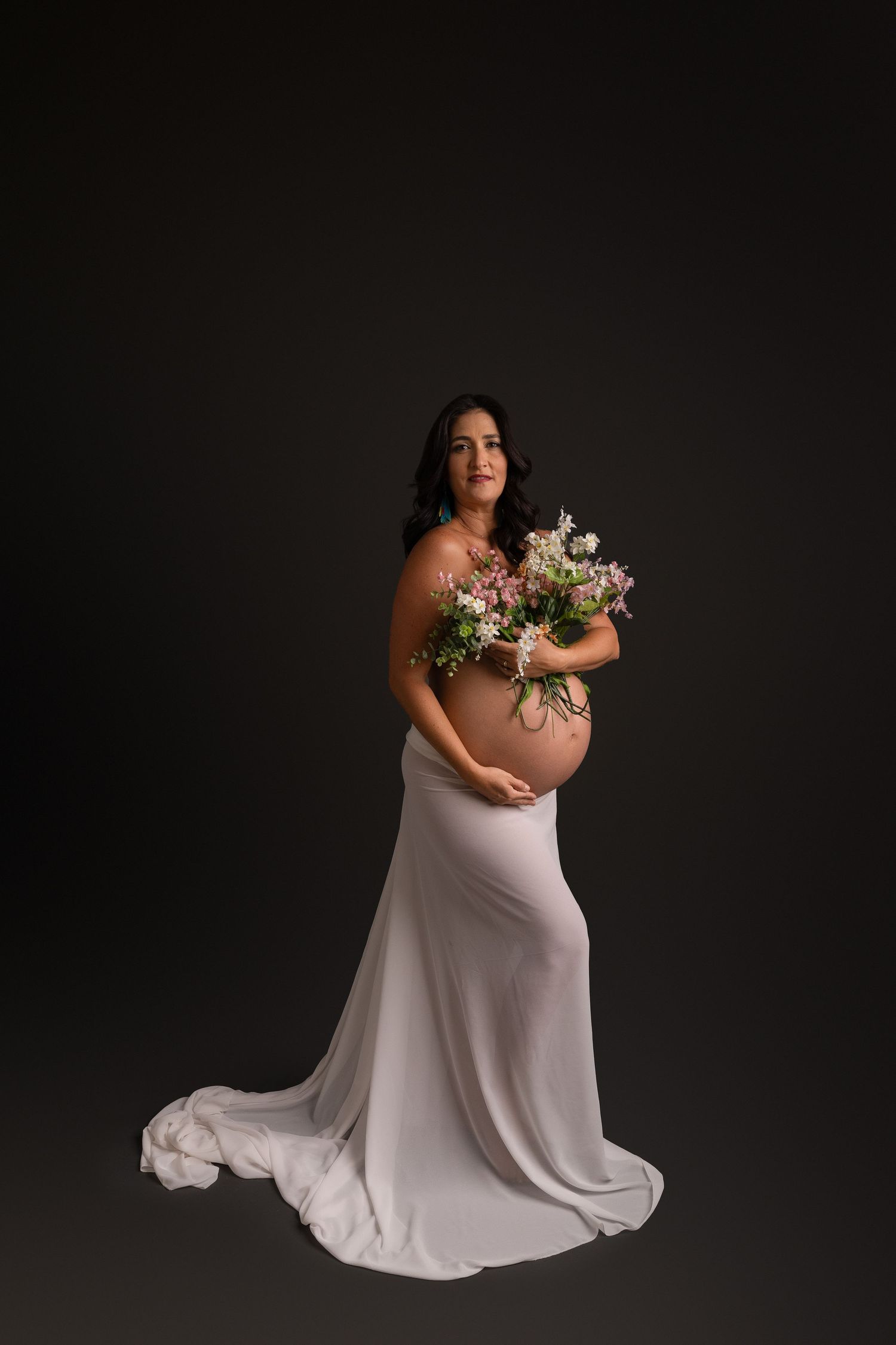 Artistic maternity photoshoot featuring white flowing fabric and floral elements against a dark background.