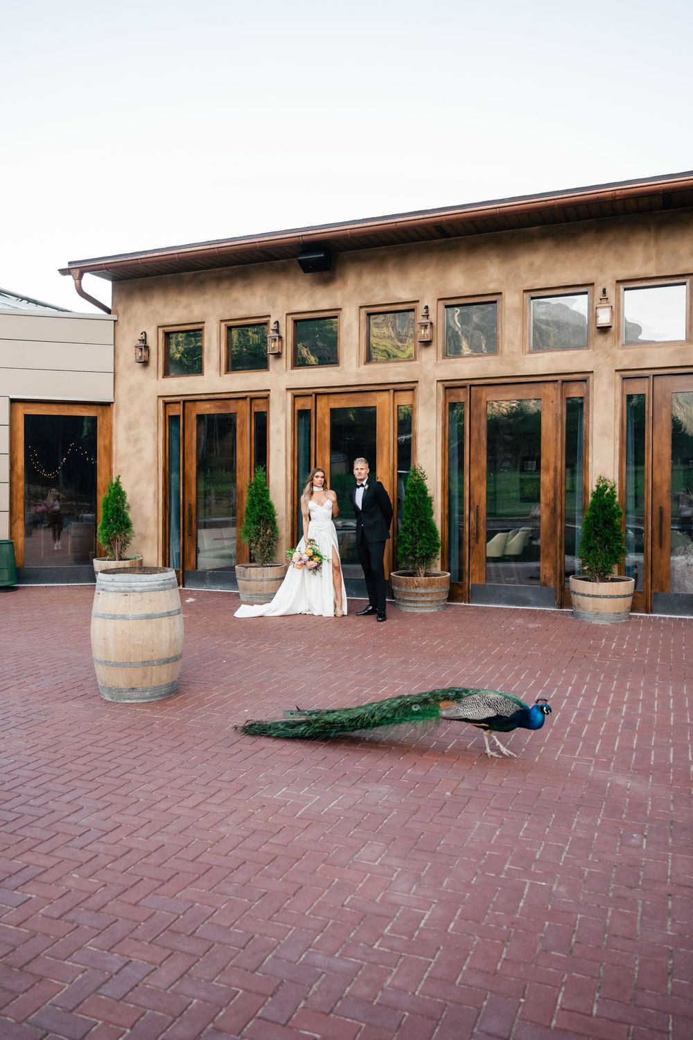 Wedding couple stands in front of rustic venue with large wooden barrel decor on brick patio in garden setting.