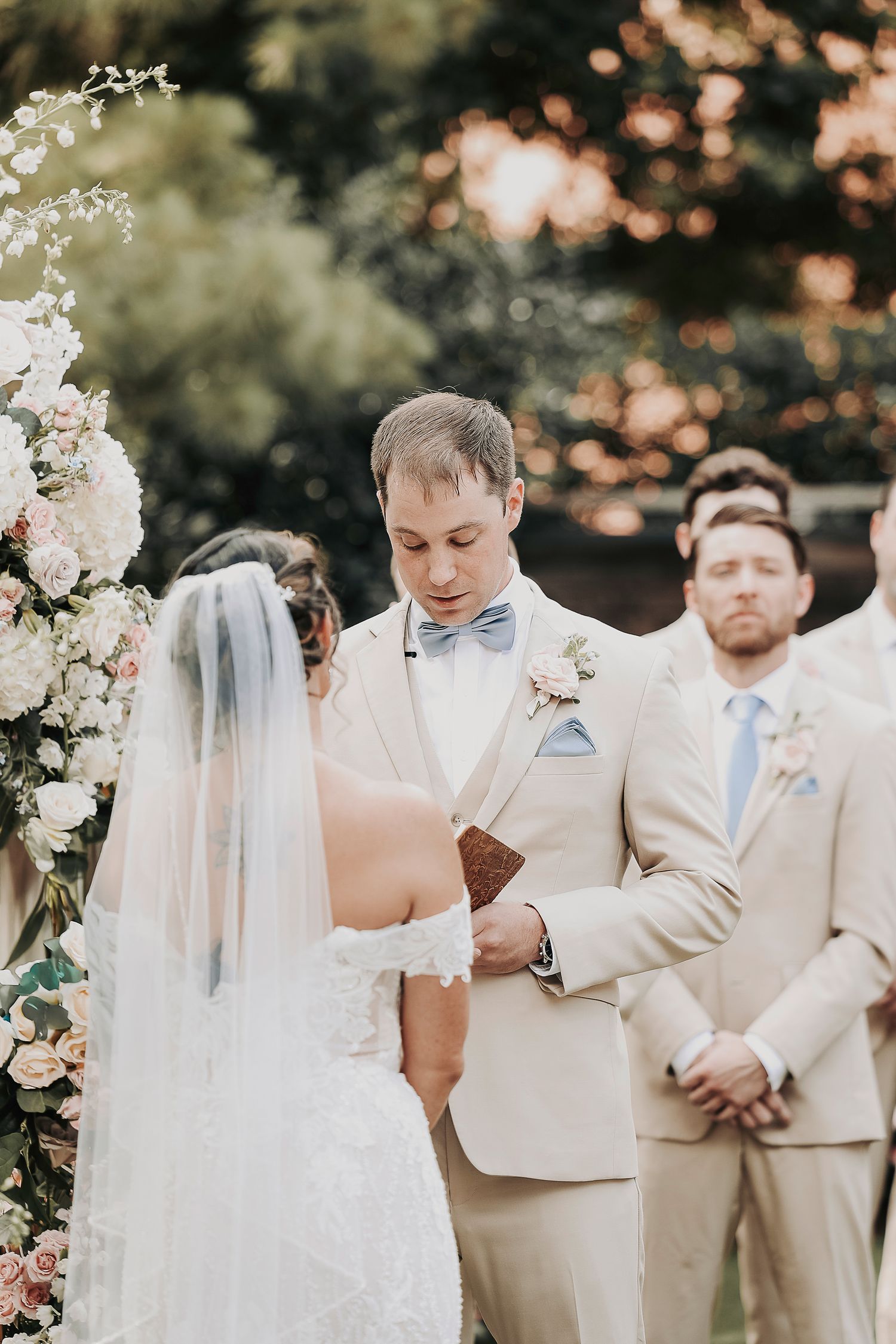 Bride and groom exchanging vows under floral arch with soft greenery and blush tones