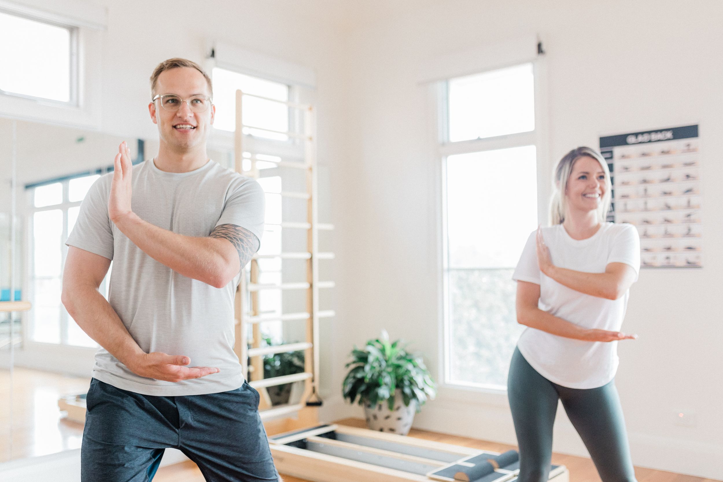 Two people exercising and stretching in a bright fitness studio with plants and windows.