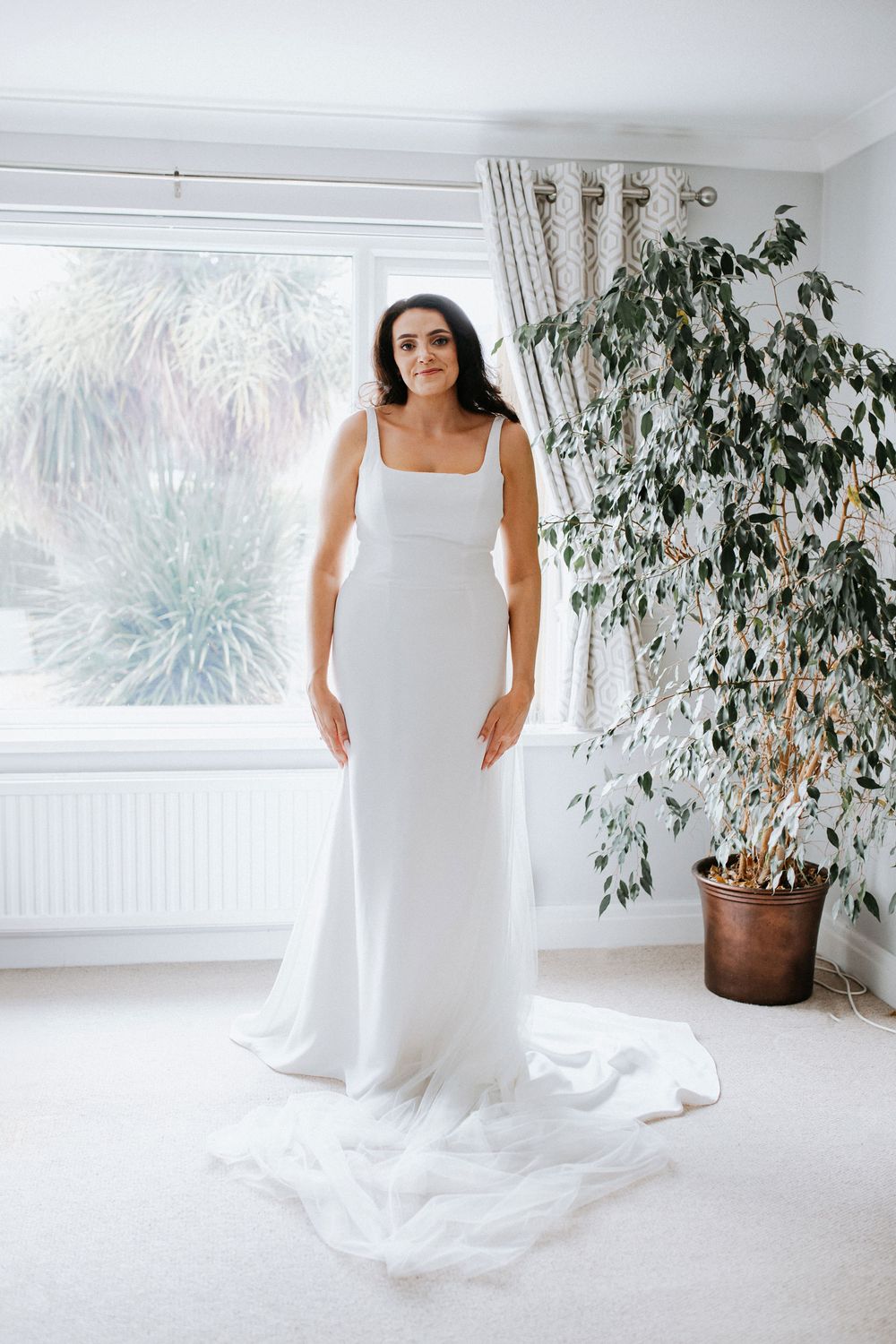 A bright indoor photoshoot featuring a flowing white dress against light walls with potted plants as natural decor.