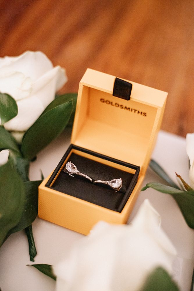 An engagement ring displayed in a gold Goldsmiths jewelry box nestled among white flowers and green leaves.