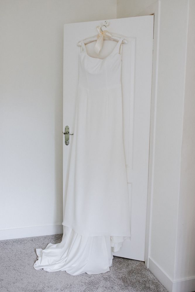 A simple white wedding dress hanging on a hanger against a white door in a minimalist setting.