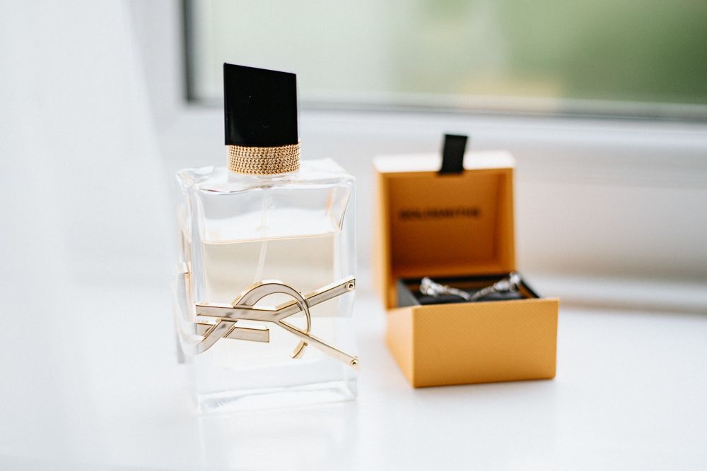 Elegant perfume bottle with gold YSL logo sits beside orange designer box on white windowsill.