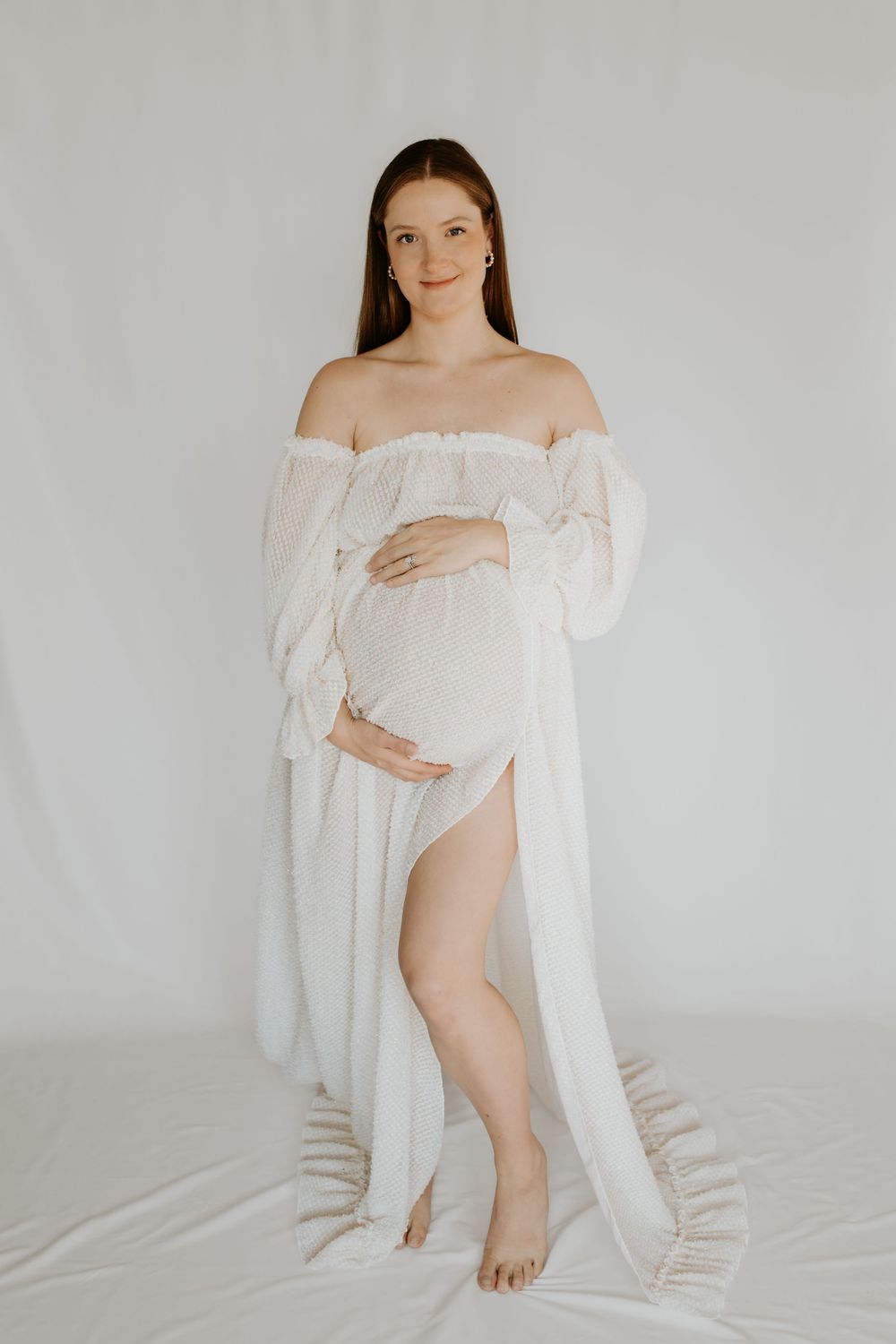 An elegant maternity portrait showing a person in a flowing white off-shoulder dress against a light background.