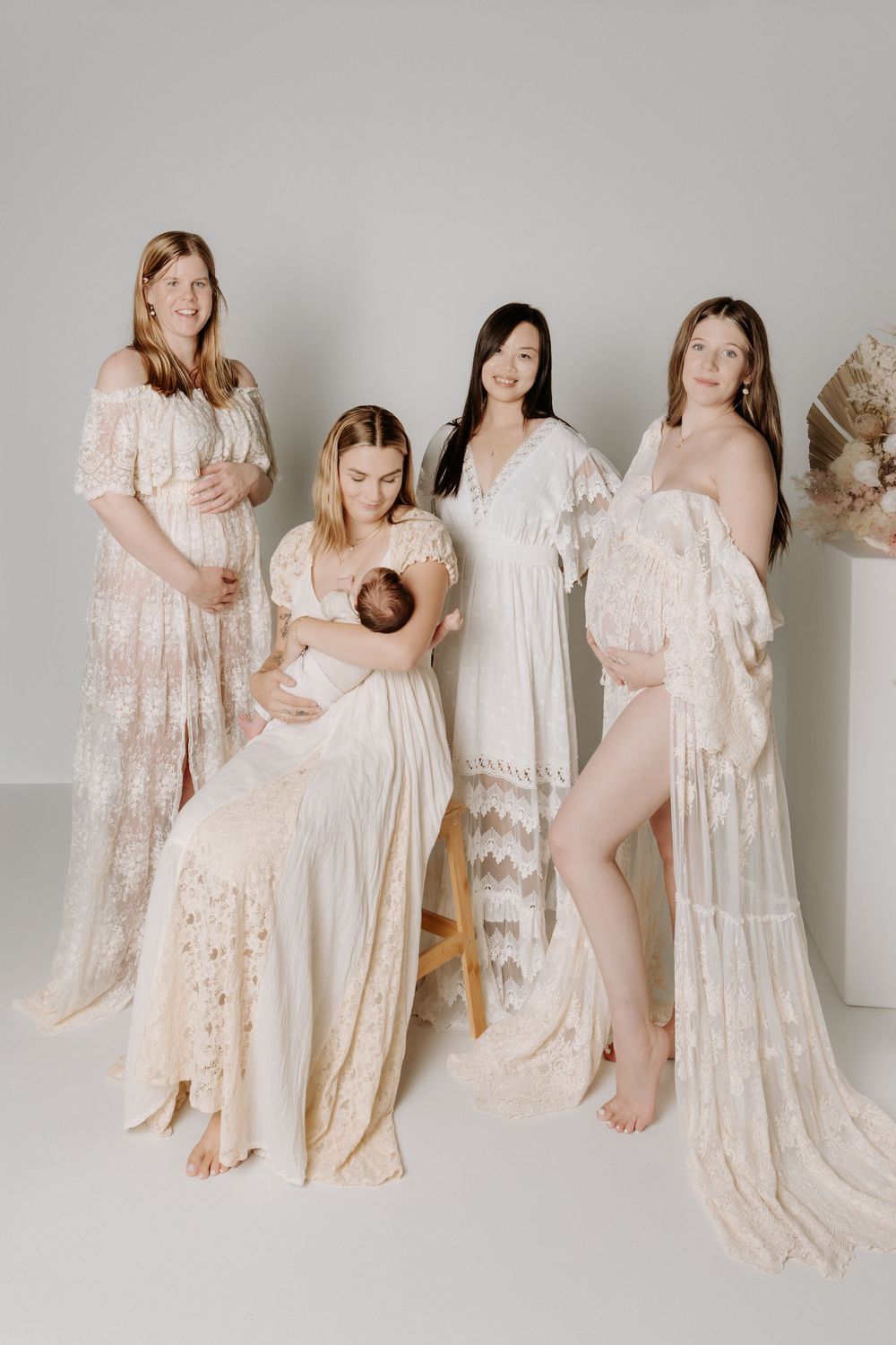 Group of expecting mothers in flowing white lace dresses pose together in an ethereal maternity photo shoot with a newborn.