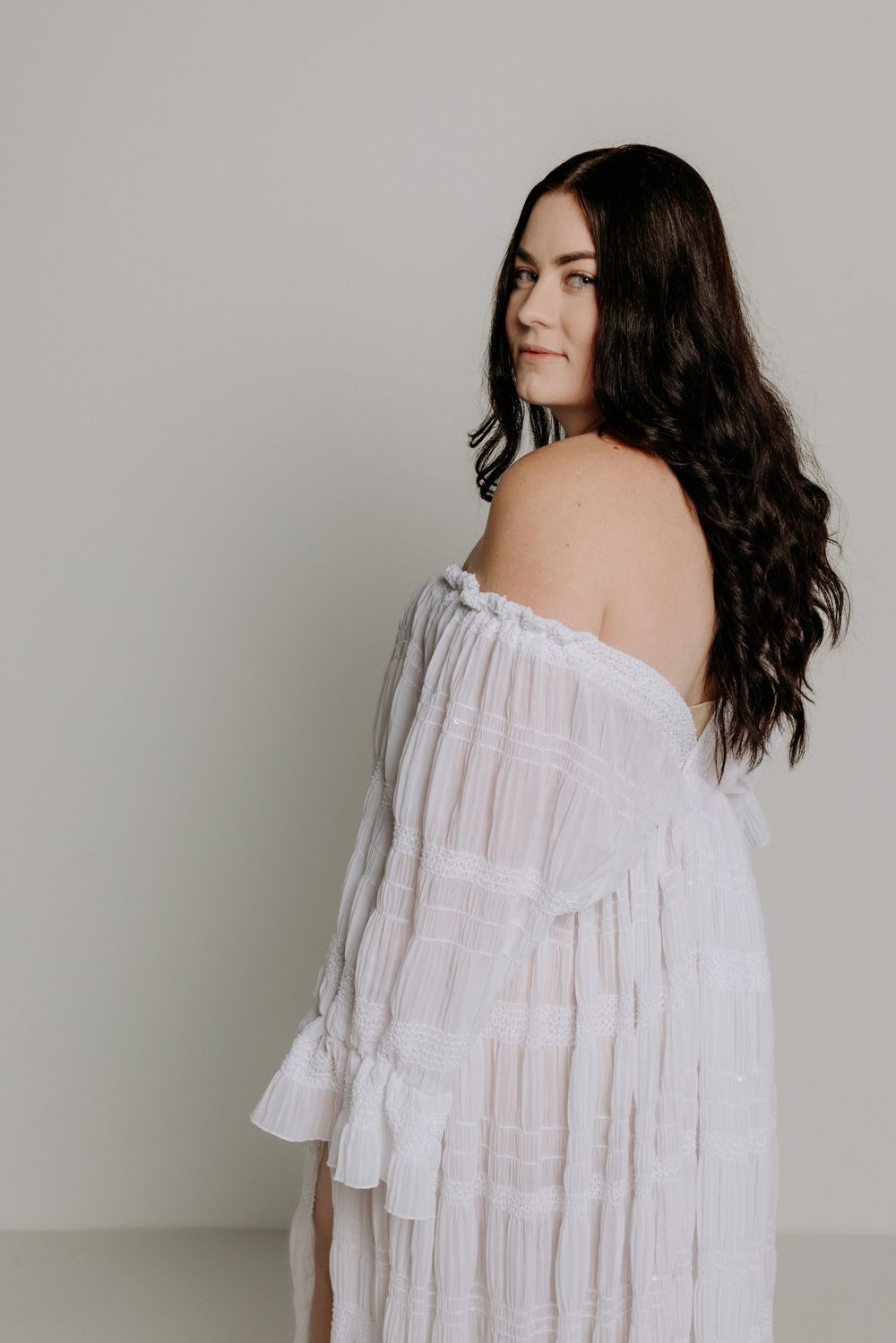 A portrait of a woman with dark wavy hair wearing a white off-shoulder dress against a plain backdrop.