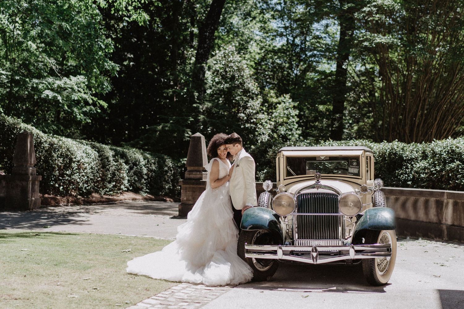Bride and groom standing beside a vintage car, smiling and enjoying their moment together.