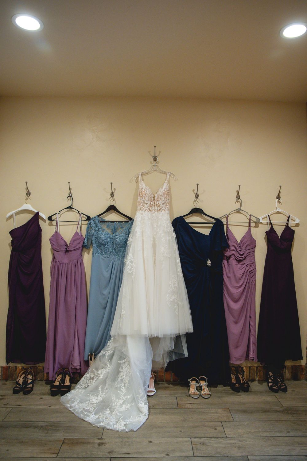A wedding dress and bridesmaid dresses in purple, mauve and navy hanging on a wall with shoes beneath them.