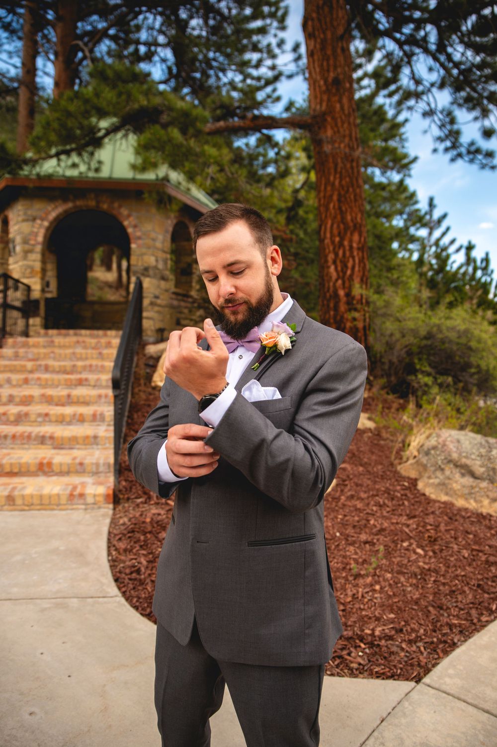 A person in a gray suit adjusts their sleeve cuff near a stone gazebo with pine trees in the background.