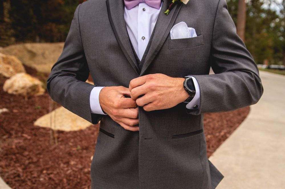 Close up details of grey wedding suit with purple bow tie, white pocket square and wristwatch against garden backdrop.