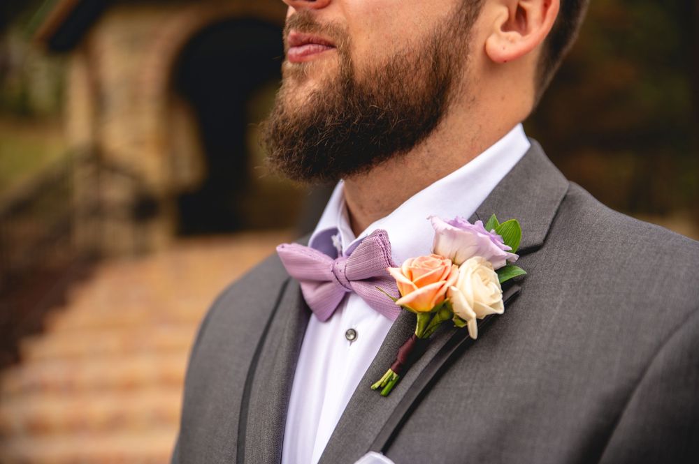 Close up of groom in grey suit with peach rose boutonniere and lavender bow tie at outdoor wedding ceremony.