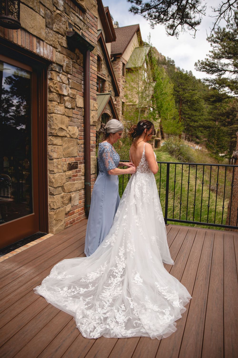 Beautiful wedding dress with intricate lace train flows across wooden deck of rustic stone mountain lodge.