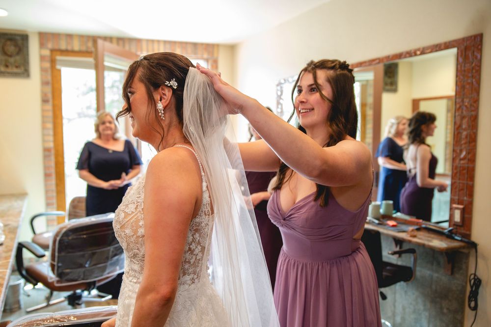A bridesmaid in a purple dress helps adjust the bride's wedding veil in a salon preparation room.