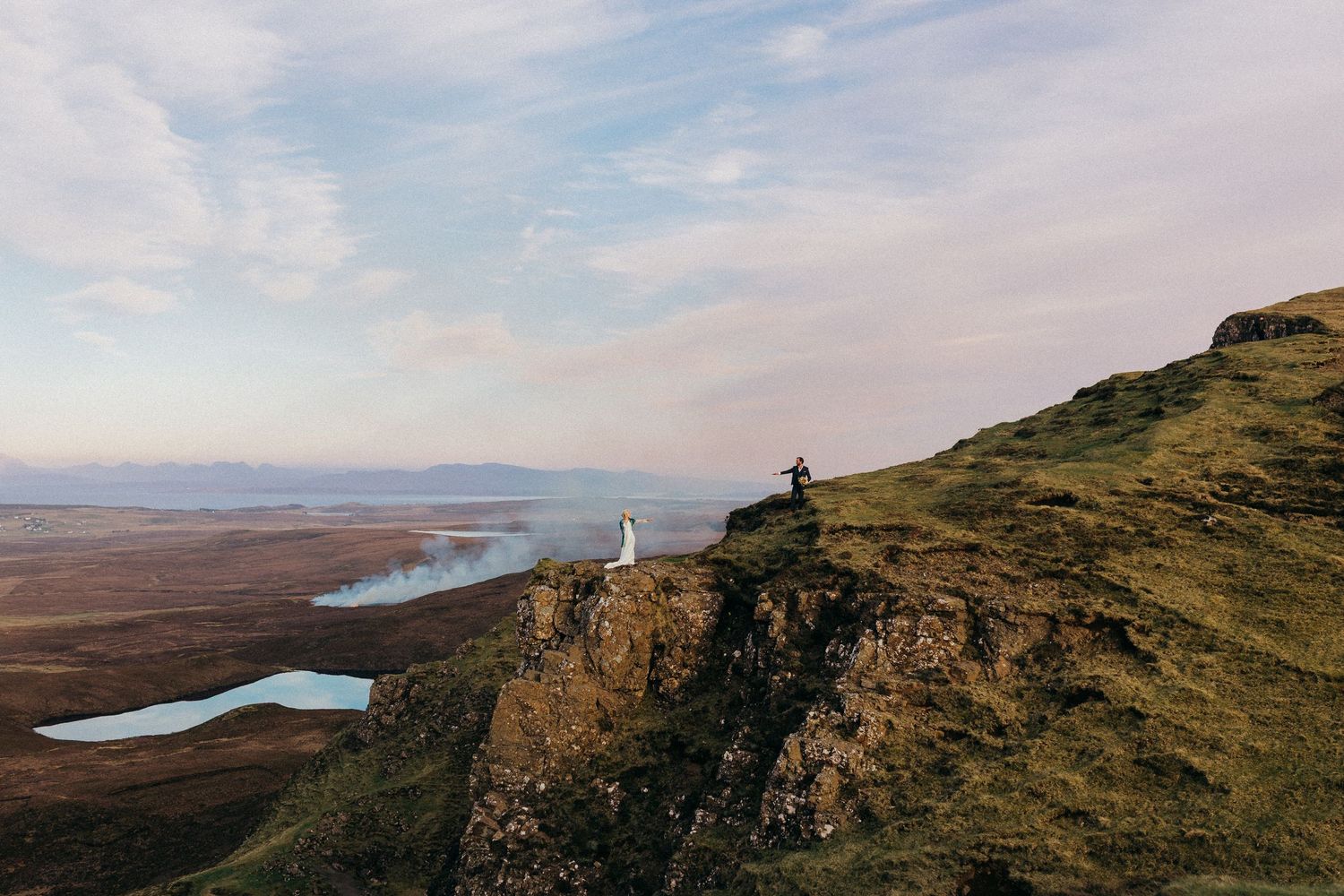 Scenic view of a person standing on cliff edge overlooking misty lakes and rolling hills in remote landscape.