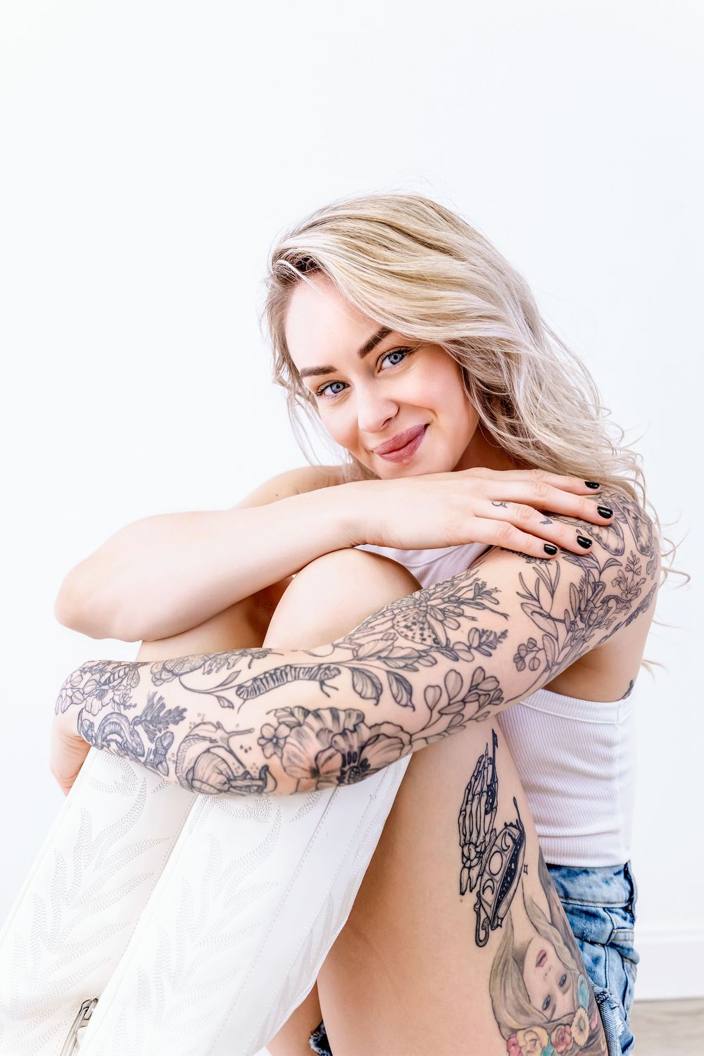 Blonde woman in white tank top showing floral sleeve tattoos while sitting in casual pose.