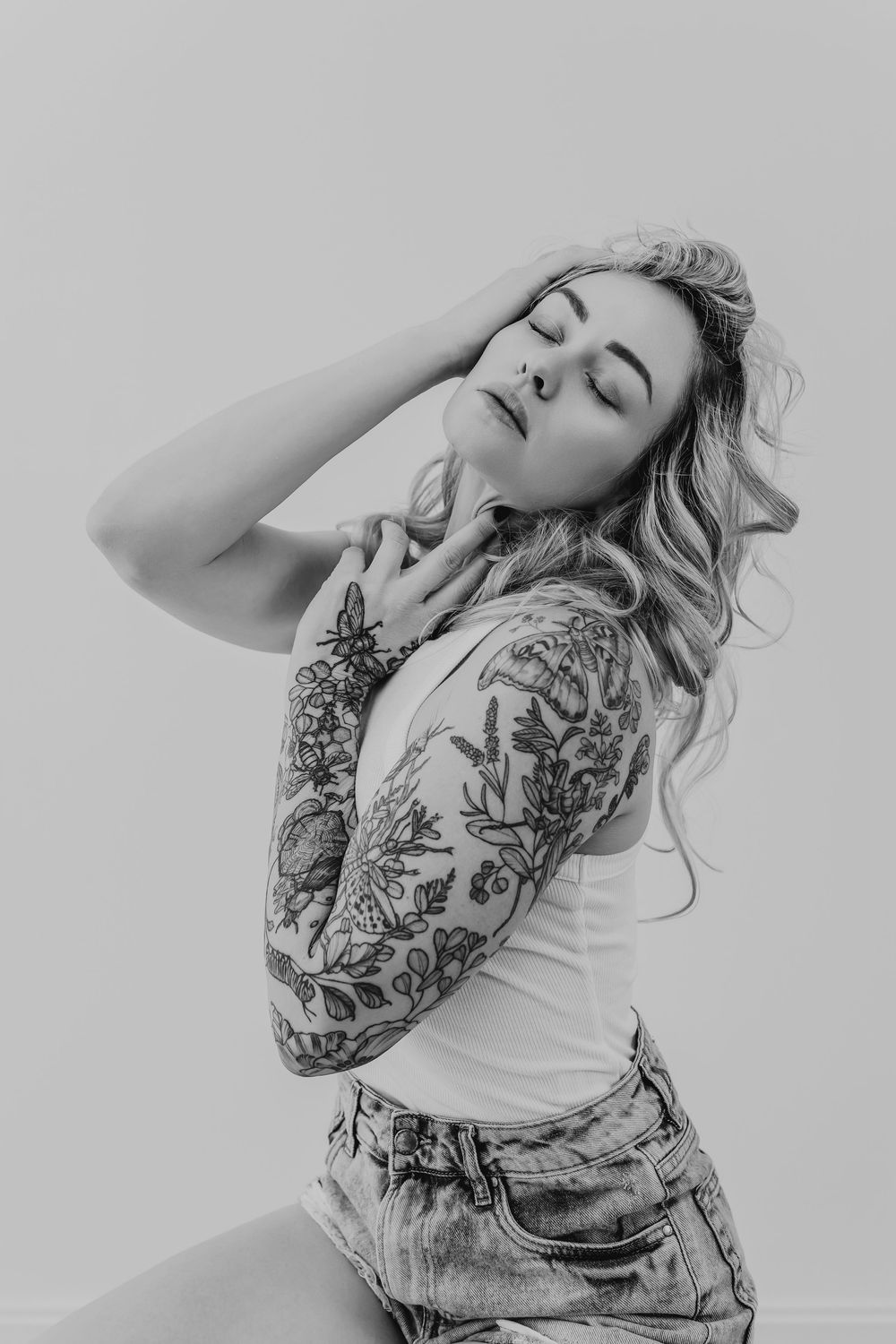 Black and white portrait featuring artistic floral sleeve tattoos and wavy blonde hair against light background.