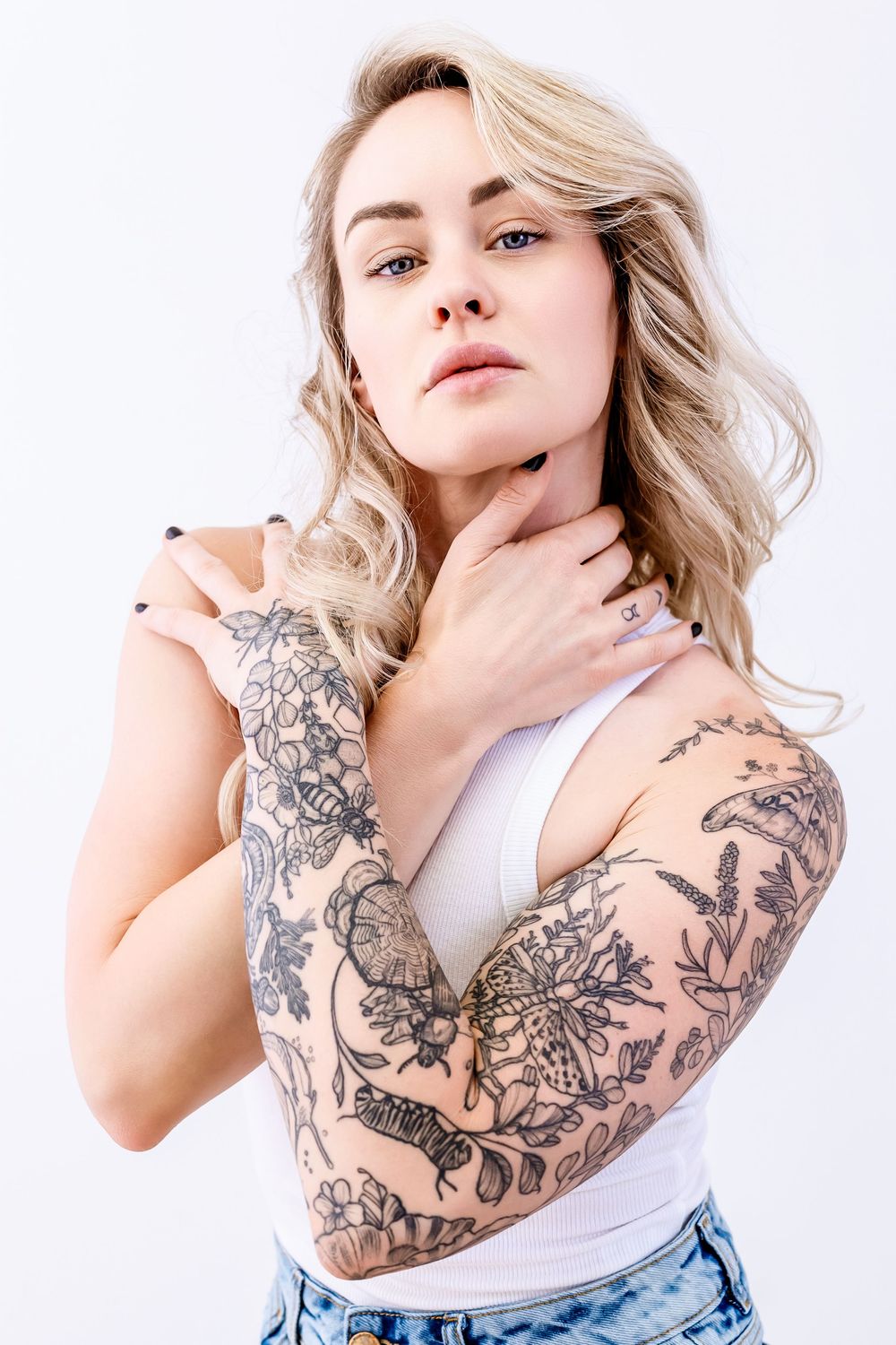 A model in a white tank top poses showing floral tattoo sleeve artwork.