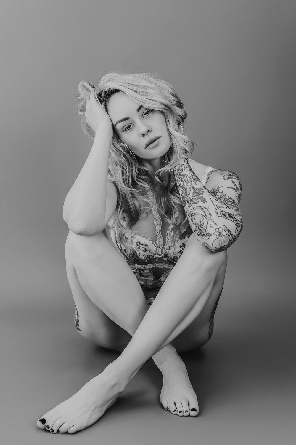 Black and white artistic portrait of a woman in casual pose sitting cross-legged on floor with wavy blonde hair.